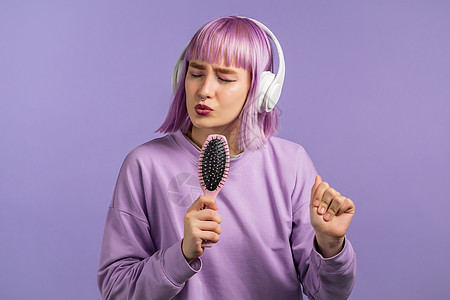 紫罗兰色发型的年轻漂亮女人用发刷或梳子唱歌跳舞 而不是在紫罗兰色墙上的麦克风 戴耳机的女孩玩得开心 听音乐 梦想成为名人图片