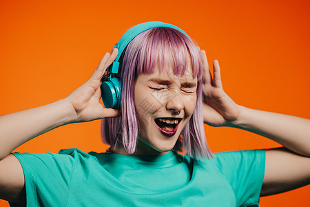 穿着染色紫色紫色头发 耳机听音乐和橙色背景歌唱的特异女性 独特的时髦女孩舞蹈成人乐趣女士自由立体声喜悦工作室享受青少年快乐图片