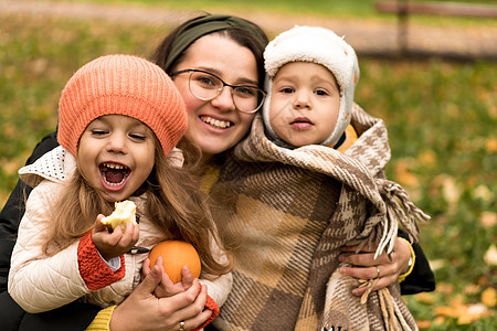 年轻的女人妈妈和小可爱的学龄前未成年儿童穿着橙色格子在黄色落叶上在秋天公园寒冷的天气里微笑着看着相机 童年 家庭 母亲 秋天的概图片