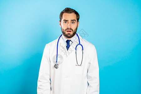 身穿专业医用白大衣的年轻失望男子的肖像 该名男子被隔离在蓝工作室背景上 有胡子和听诊器的健忘医生图片