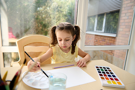 顶端风景 穿黄色T恤和两匹马尾辫的学龄前女孩在家美术课上享受水彩画图片