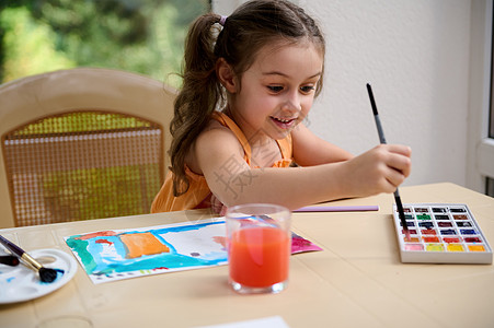 可爱的小女孩坐在桌边 一边用画笔混合颜料 一边在美术课上学习绘画图片