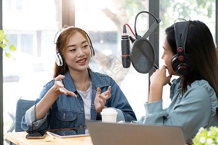 微笑着两个亚洲年轻女性 男性电台主持人戴着耳机 在谈话时使用麦克风 谈话 在演播室的广播中一起录制播客 制作录音音频概念的技术嗓图片