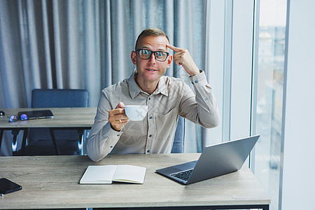 戴眼镜的自由职业者在笔记本电脑上工作并喝咖啡 经理坐在办公室的桌子旁 在笔记本电脑上工作 自由职业者工作场所 远程工作的员工男性图片