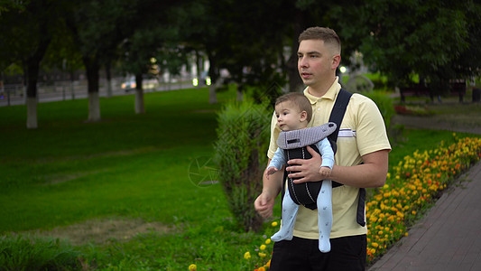 一个年幼的父亲在公园里带着孩子走 新生儿子在袋鼠背包里图片