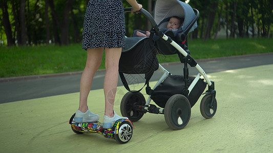 年轻母亲站在公园的悬浮板上时携带婴儿车 在公园与孩子一起走现代路程图片