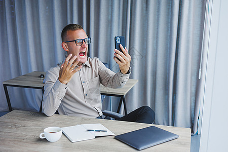 一位戴眼镜的年轻迷人男性自由职业者坐在办公室里 看着电话 帅气的博主坐在办公室的木桌旁 拿着笔记本电脑在电话上写想法阅读商业职场图片