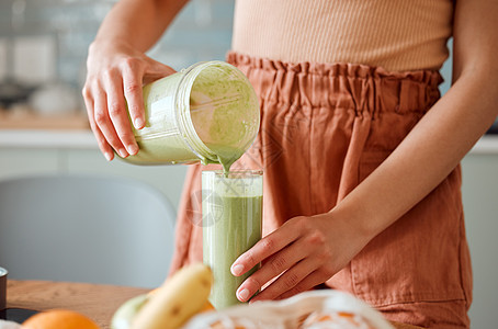 女人从柜台上的搅拌罐中将健康的冰沙倒入玻璃杯中以排毒 女性在她的厨房里用蔬菜和消耗品制作新鲜的绿色果汁 以实现健康的生活方式图片