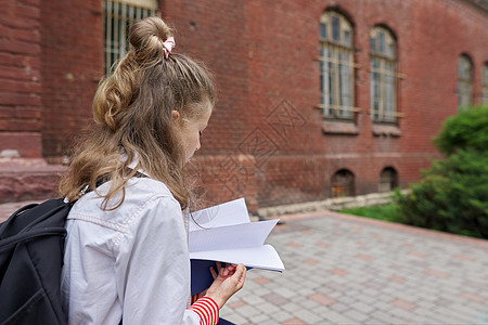 在学校院子里坐着背背包的女童阅读笔记本 复制空间孩子快乐青年学习小学生想像力女性成功学生童年图片