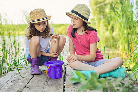 女孩坐在木码头上 在桶里抓水蜗牛假期池塘快乐闲暇游戏孩子享受友谊乐趣童年图片