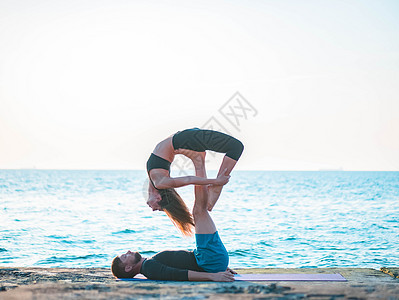 Acroyoga概念 两个运动者在自然日出的背景下对着做瑜伽 美丽的年轻夫妇在海边做伸展锻炼女士力量朋友们男人讲师训练女性男性平图片