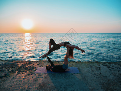 Acroyoga概念 两个运动者在自然日出的背景下对着做瑜伽 美丽的年轻夫妇在海边做伸展锻炼冒充女士花园力量海滩训练杂技女性男人图片