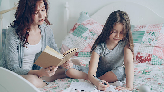 年轻的妈妈帮她可爱的小女儿做小学作业 爱妈妈在家里坐在床上看书 女孩在抄写本上写笔记图片