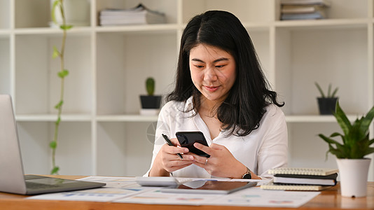 微笑的亚洲女上班族打短信 在手机上在线聊天人士报告女士消息电子邮件生意电脑冲浪技术会计图片