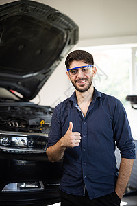 年轻的白种人留着胡子的英俊男子汽车修理工或经理的肖像 他戴着护目镜 对着镜头微笑 在车库里竖起大拇指 站立在汽车服务的快乐的人图片