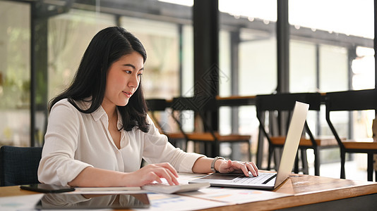 亚洲女性办公室女职员在她的笔记本电脑上查询在线信息 并在木制桌上使用计算器图片