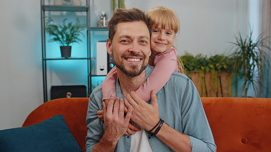 微笑的小孩儿女儿拥抱她的年轻父亲 看镜头 在家沙发上甜蜜的温柔爱图片