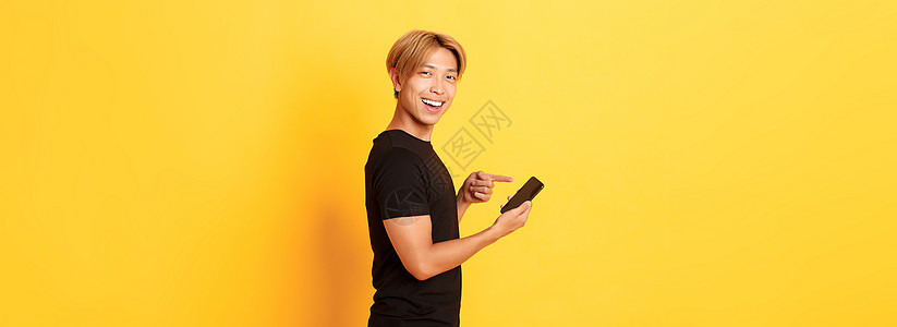 微笑的英俊的亚洲人 站在侧写和对智能手机指手指脚 推荐应用程序 站立黄色背景促销广告潮人理发男性职业情感购物技术互联网图片