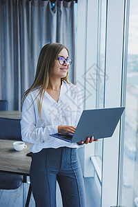 一名年轻 微笑着的女商务人士站在办公室窗边 手拿着笔记本电脑 她戴着眼镜和白衬衫 当女经理生意人会议工作男人教练团队合作人伙伴商图片