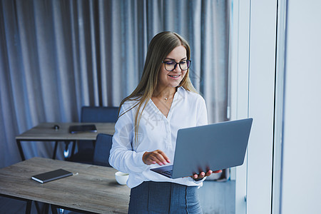 一名年轻 微笑着的女商务人士站在办公室窗边 手拿着笔记本电脑 她戴着眼镜和白衬衫 当女经理会议互联网人士管理人员支持生意人业务男图片