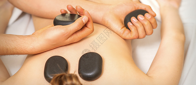 女性背部用按摩师的手进行热石按摩 在温泉沙龙中握着黑色按摩石头中心女士药品横幅说谎皮肤身体压力治疗治疗师图片