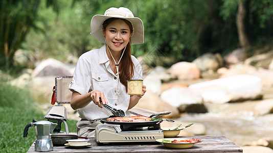 在河岸附近露营时 在便携式燃气炉上喝咖啡和烧烤bbq的热心女旅行者图片