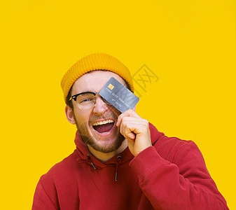 手里拿着信用卡或借记卡的年轻人开心地笑着捂住眼睛准备花钱 穿着红色连帽衫的年轻人 拿着银行卡模型 背景是黄色的 购物理念图片