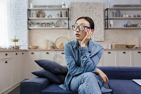 年轻漂亮的亚洲女人坐在家里客厅的沙发上 戴着眼镜思考图片