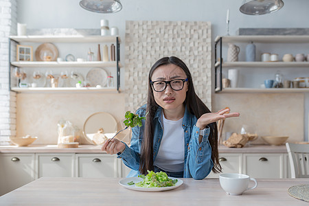 年轻不满意的亚洲妇女在家里吃沙拉和看摄影机的肖像图片