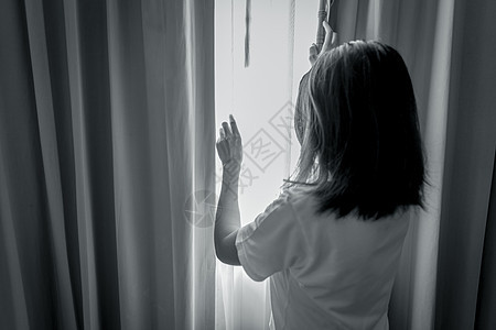 女人拿着窗帘从卧室往外看的后视图 沮丧和强调的女人 世界精神卫生日概念 心理健康意识 在黑暗的房间里体贴的女人 不幸的女人背景图片