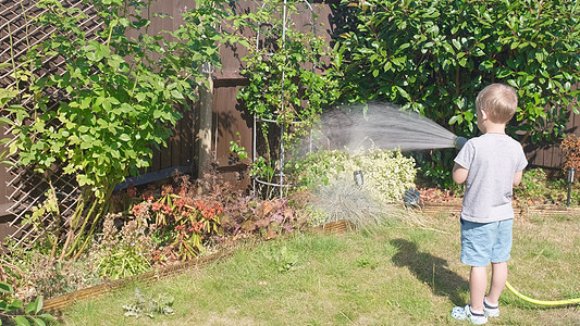 有趣的小男孩在花园房屋后院浇灌草坪植物 可爱的孩子在炎热的阳光明媚的夏天在户外玩灌溉软管 孩子们帮忙做家务 孩子们的活动 童年太图片