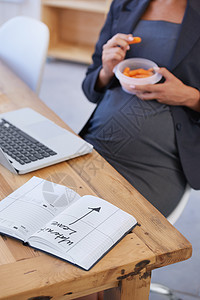 期待她的产假 一位正在吃零食的孕妇躺在办公桌上 上面写着详细说明产假的笔记本图片