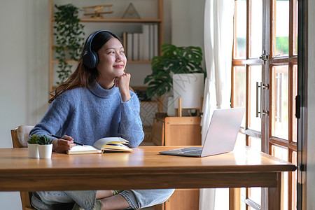 音乐老师戴着耳机的微笑女孩坐在办公桌前 看着外面 用笔记本电脑在线学习 喜欢戴耳机的聪明年轻女性参加网络课程或练习使用计算机 远程教育理背景
