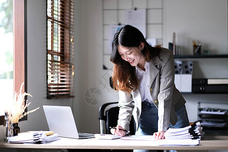 在办公室使用计算器和笔记本电脑的亚洲女商务人士快乐地站在那里工作商业互联网商务微笑女士桌子秘书工人人士图片