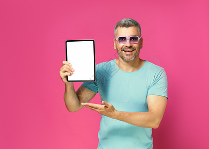 推出一个大提议 显示数字平板白屏幕的英俊男子观看照相机时看到穿蓝衬衫的相片和粉红色背景孤立的太阳镜 App广告图片