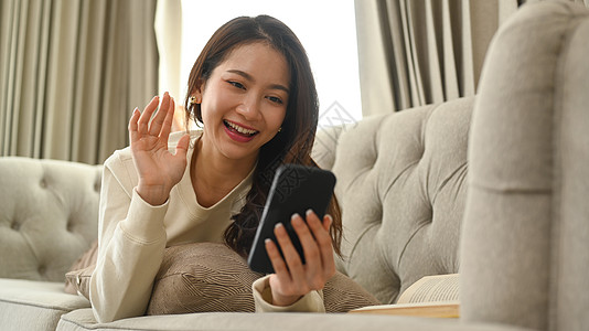 快乐的年轻女子挥手 通过智能电话进行视频通话 同时在家睡沙发上休息图片