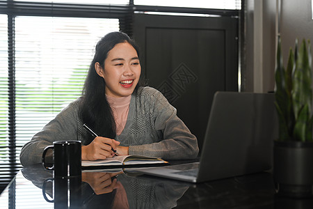 微笑的亚洲人看着笔记本电脑屏幕 观看在线讲座并在笔记本上做笔记图片
