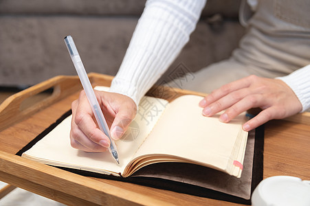 一位身穿工装裤和毛衣的年轻女性坐在木制托盘桌上的皮革记事本上用黑色凝胶笔书写的特写 她坐在家里沙发旁的地板上作家学习教育桌子笔记图片
