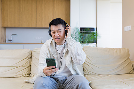 年轻开朗的亚洲人坐在家里的沙发上 戴着耳机听音乐 一个人独自坐在沙发上感觉很好的生活方式 拿着电话的快乐男性在网上收听他最喜欢的图片