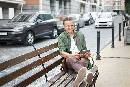 自由职业者快乐工作的人使用数字平板电脑工作 坐在前办公室大楼户外的长凳上 身着绿色散装衬衫和肩膀袋 穿着便衣图片