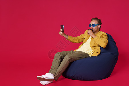 快乐英俊的年轻人 30 多岁使用数字平板电脑从纸杯喝咖啡自拍穿着休闲牛仔装坐在袋椅上隔离在红色背景 咖啡店经营理念互联网男人椅子图片