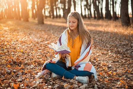 年轻可爱的女学生坐在秋天森林的格子上 阅读一本书图片