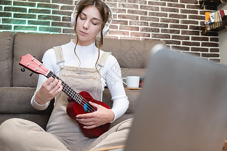 穿着休闲服的漂亮年轻时髦女孩坐在家里客厅的地板上学习弹尤克里里吉他 爱好休闲时间概念房间闲暇女士电脑歌曲乐器教育潮人艺术家幸福图片