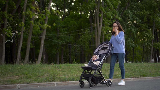 妈妈推着婴儿车带着婴儿走路 边走边吃汉堡 一位戴眼镜的年轻女子和她的儿子推着婴儿车 在公园里吃着汉堡图片