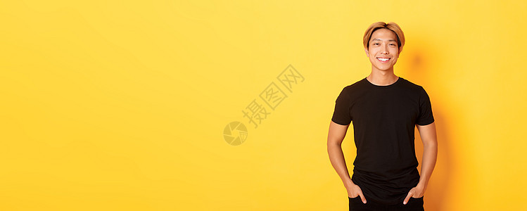 穿着黑色T恤 站立快乐 黄色背景的迷人笑笑着的亚洲男人成人理发发型促销购物职业广告金发情感学生图片