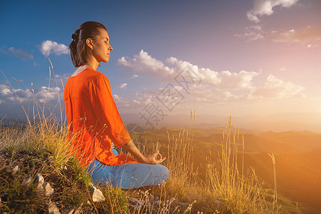 年轻的天主教女青年沉思于莲花的位置 在山上的草地上 靠近日落的悬崖边冥想训练瑜伽闲暇呼吸山脉阳光咒语女性膝盖图片