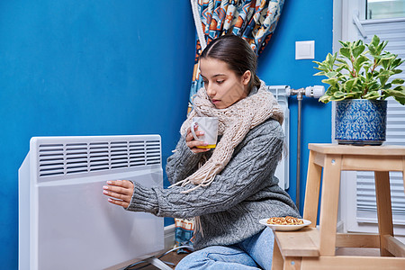 穿着毛衣围巾的少女在电热暖气散热器附近升温 喝热饮生活房子散热器气候温暖季节器具温度衣服舒适度图片