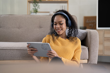 坐在客厅使用数码平板电脑的无线耳机中 年轻笑着的黑人女人的肖像图片