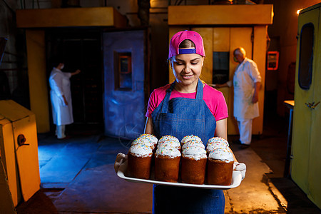 在面包店里 女孩面包师拿着一个托盘 上面放着热糕点 她穿着牛仔连衣裙和帽子 烘焙产品的生产 架上新鲜的脆皮糕点烤箱职业商业厨房制图片