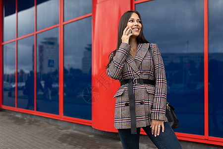 一名穿着秋大衣的年轻女子 在红色蓝色建筑外表的背景下 用手机说话 (笑声)图片